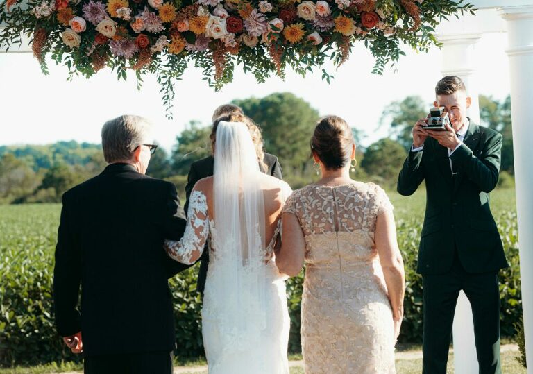 groom with polaroid of bride coming down the aisle green suite and floral canopy