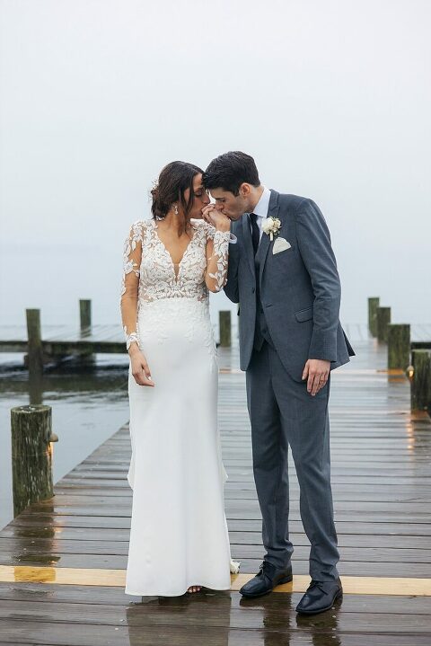 sweet hand kiss bride and groom lace dress pendant earrings and grey suite on pier at Herrington on the Bay Annapolis waterfront wedding